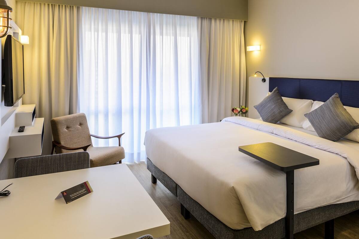 Une chambre d’hôtel comprenant un lit, un bureau, une chaise, une table de chevet et une grande fenêtre avec des rideaux.