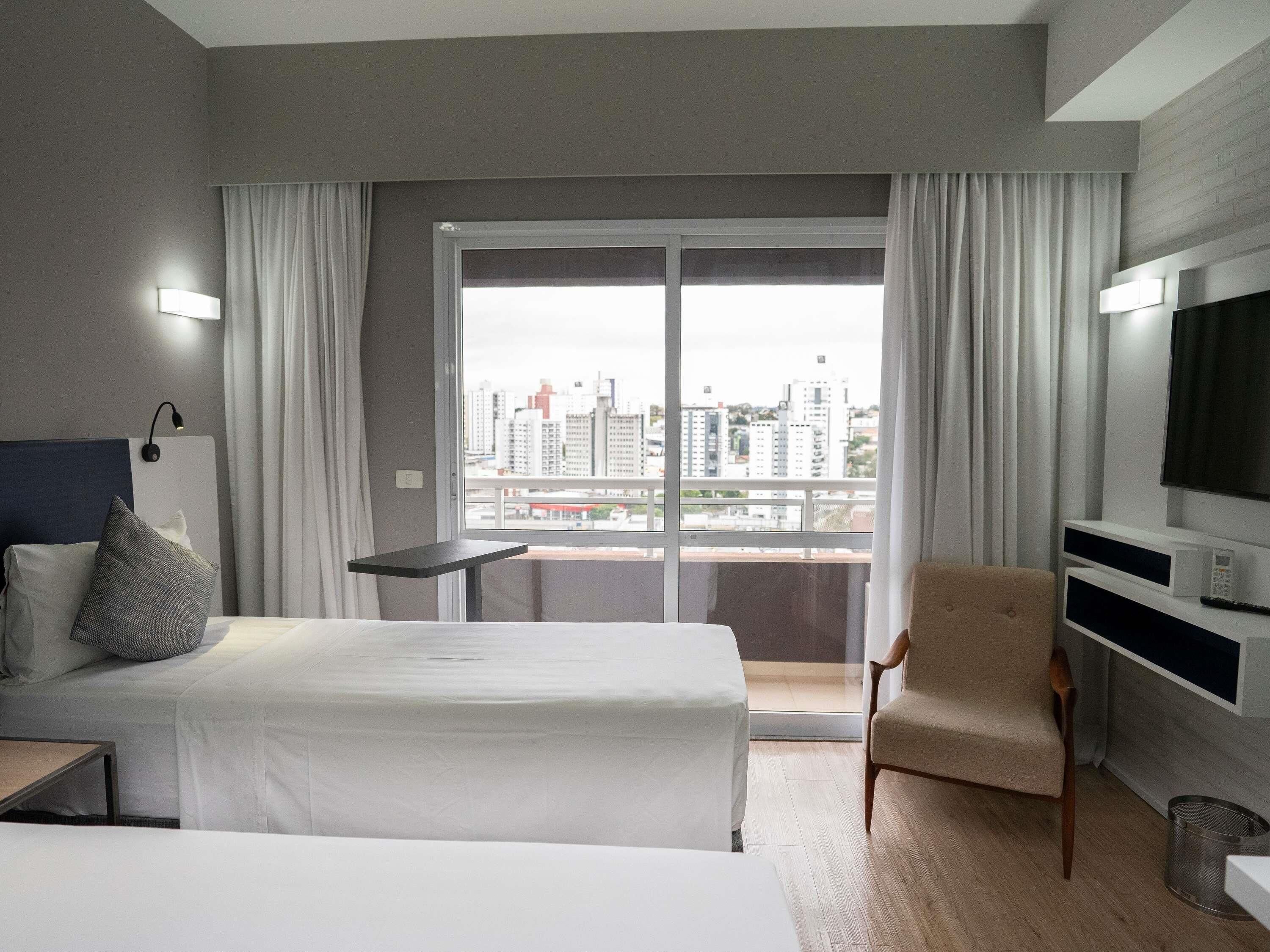 A hotel room with a bed, a chair, a desk, a TV, and a view of the city through a large window.
