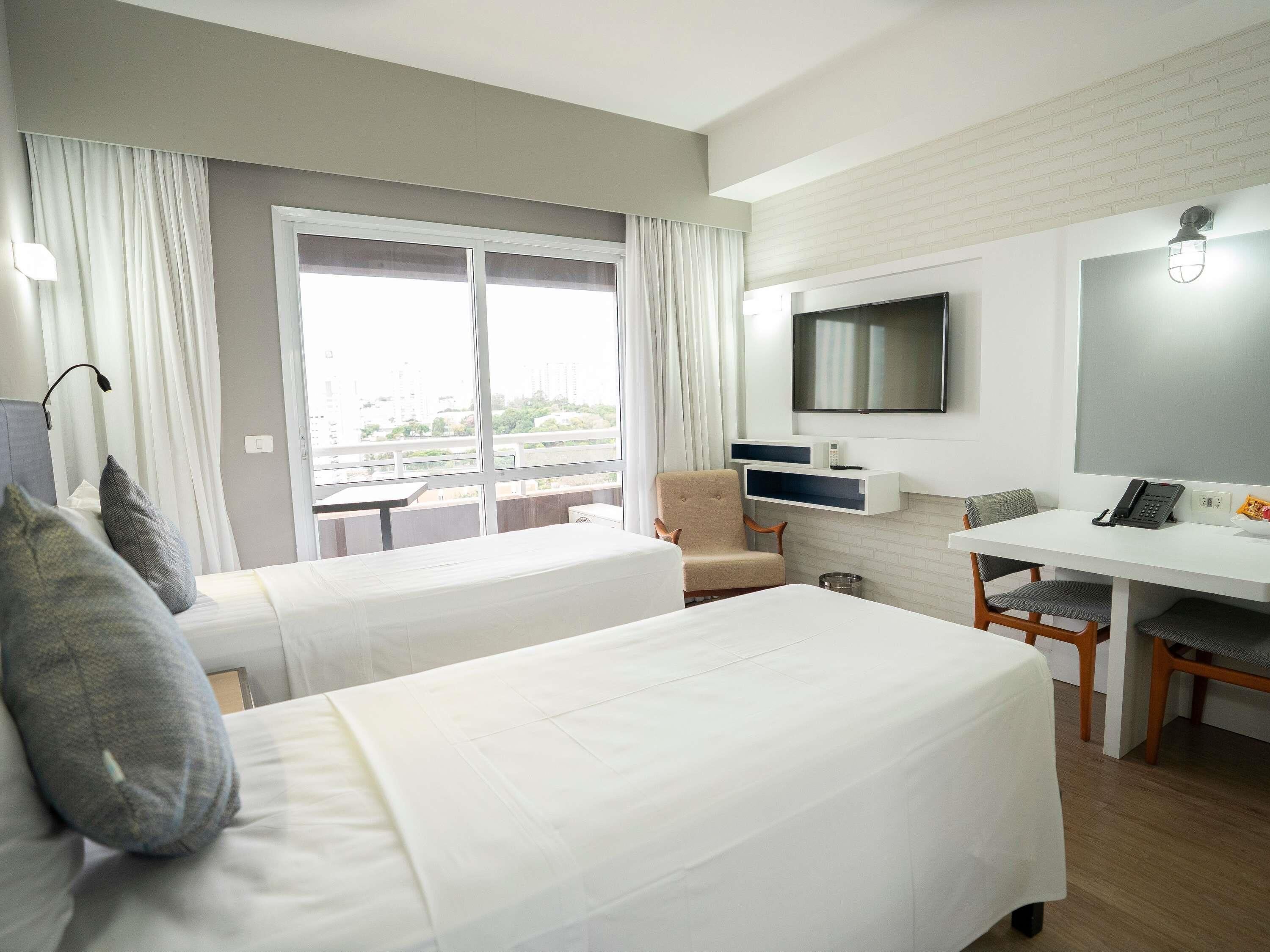 Een hotelkamer met twee bedden, een bureau, een stoel, een televisie en een balkon met gordijnen.