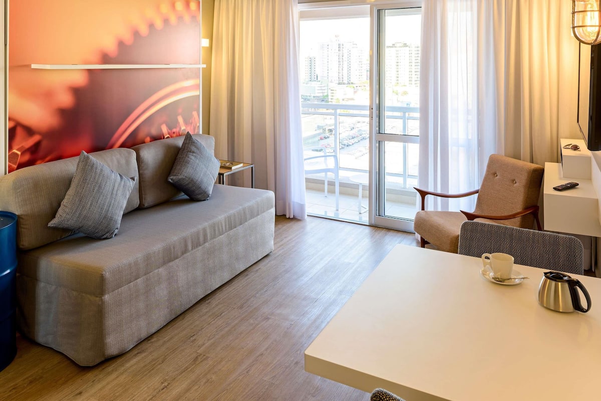 Une chambre d’hôtel avec un canapé, une table avec une tasse et une bouilloire, une chaise et une vue sur les bâtiments par la fenêtre.