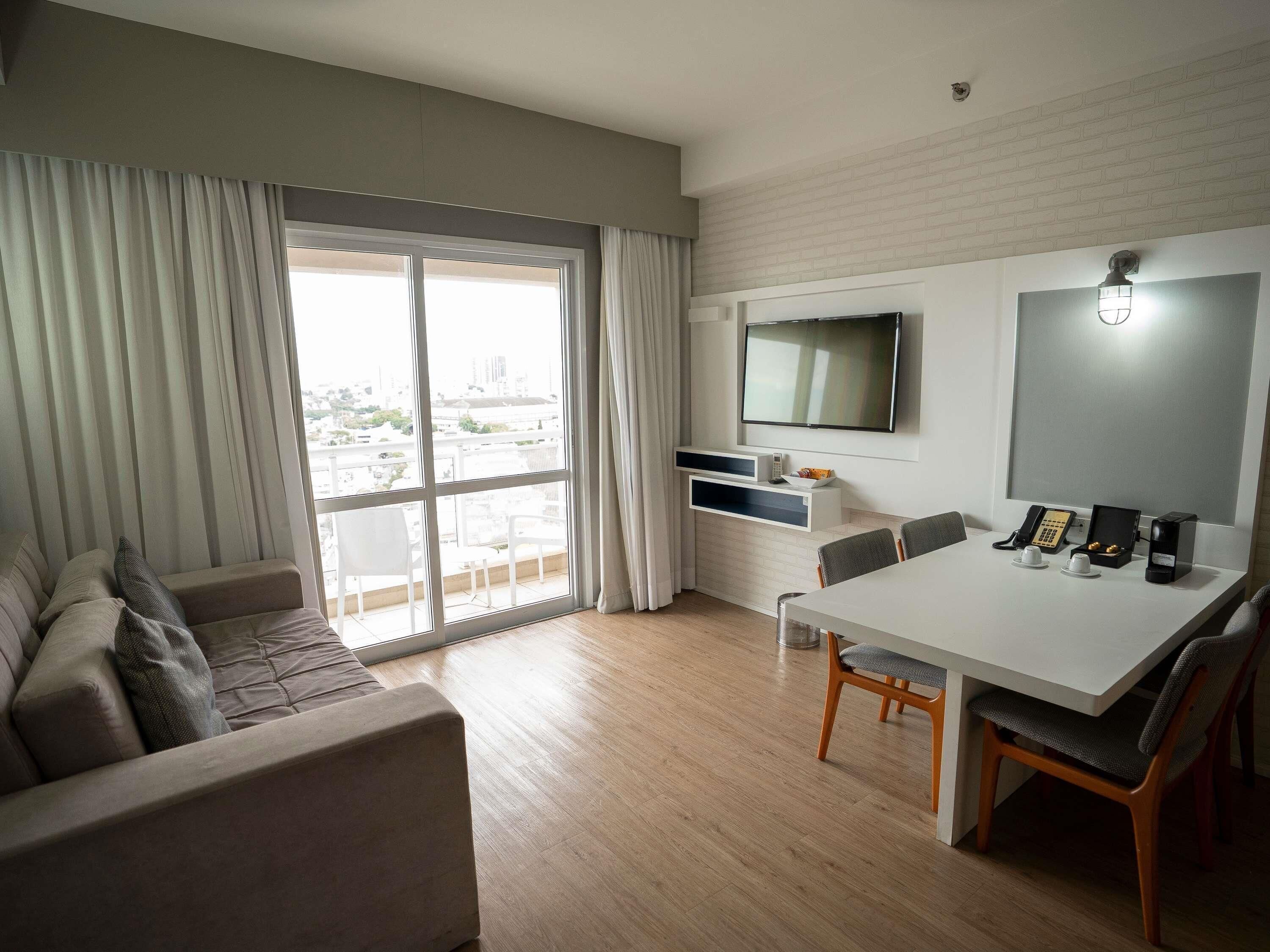 A modern hotel room with a sofa, dining table, chairs, a TV, a phone, and a coffee maker.