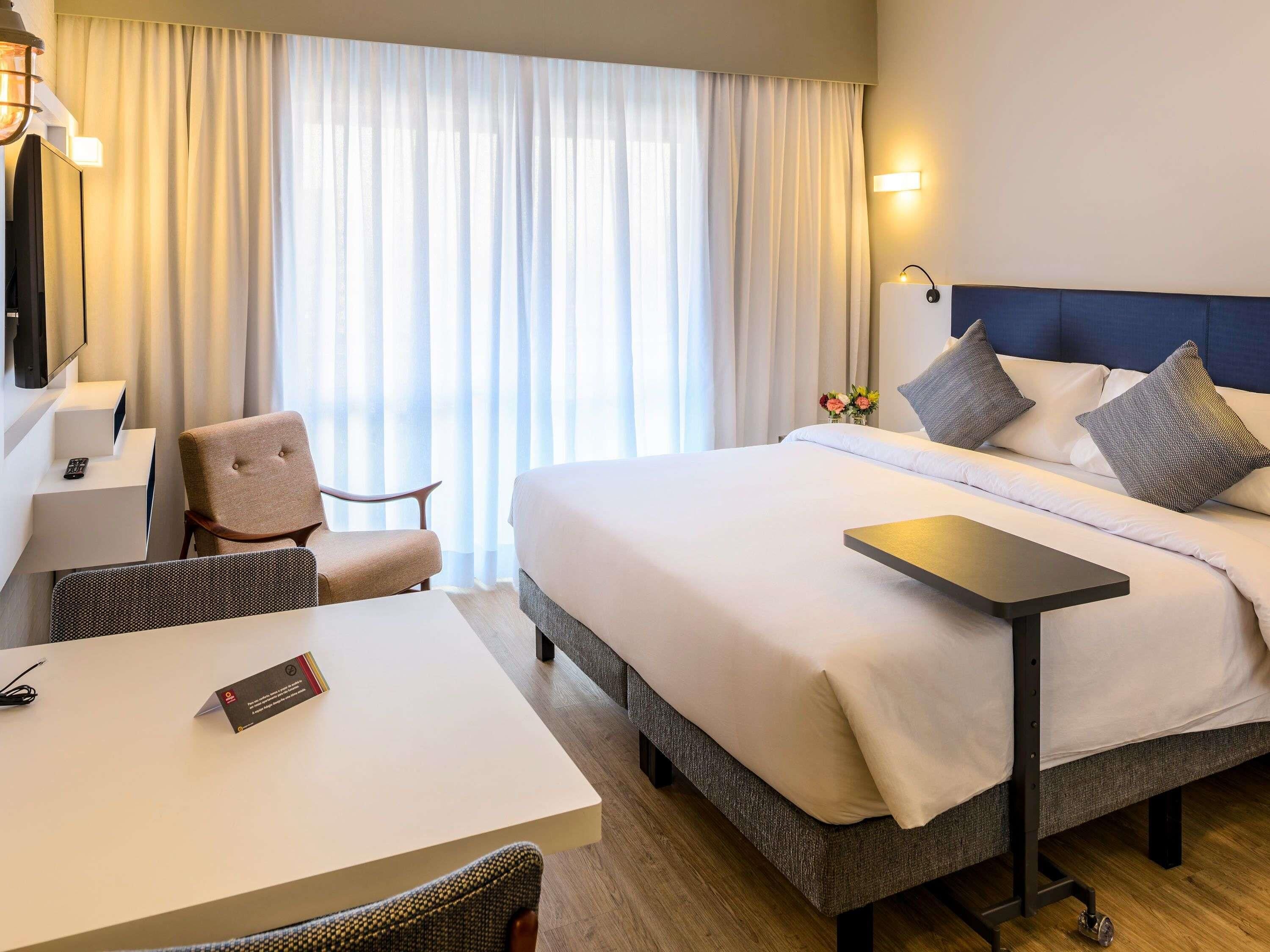 Une chambre d’hôtel avec un lit, un bureau, une chaise, une télévision et une fenêtre avec des rideaux.