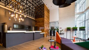 Een moderne lobby met een houten receptiebalie, hangende verlichtingsarmaturen en een zithoek met een groen tapijt.