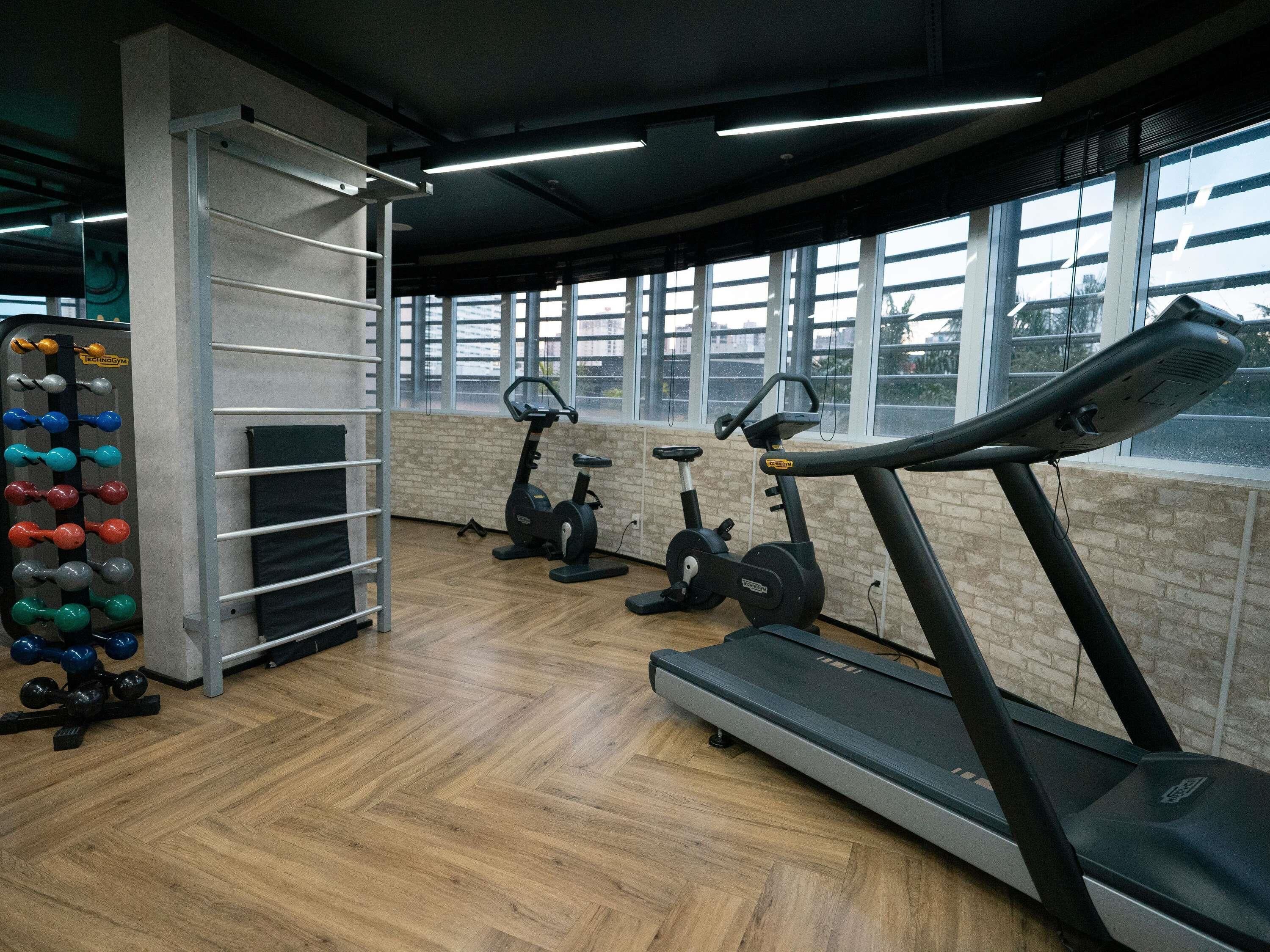Un gimnasio con equipo de ejercicio, piso de madera y ventanas amplias.