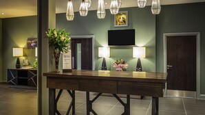 Un espace d’accueil avec un comptoir en bois, deux lampes fixées au mur, une plante en pot et un tableau encadré au-dessus d’une porte.