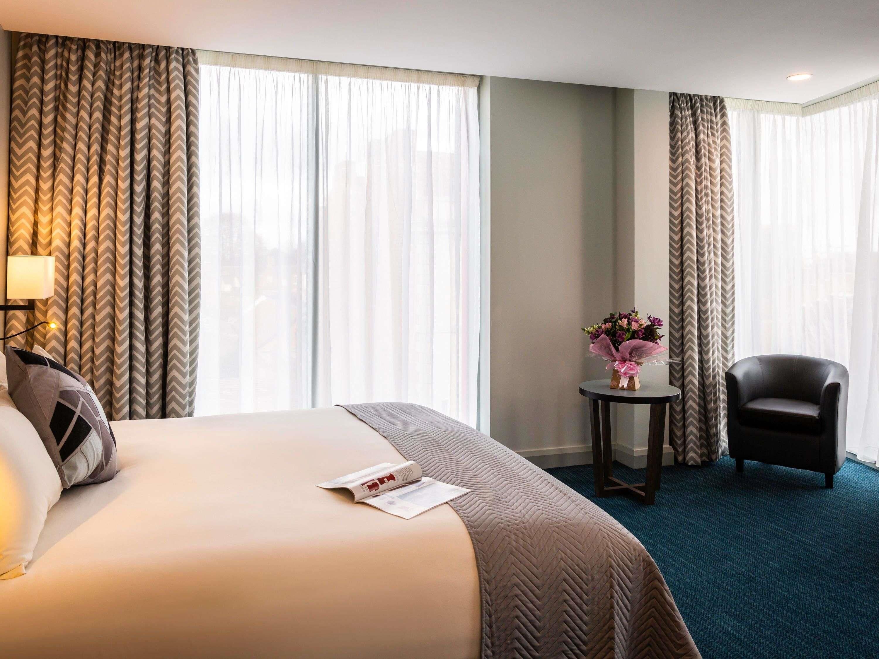 A hotel room with a bed, a chair, a small table with a vase of flowers, and a lamp.