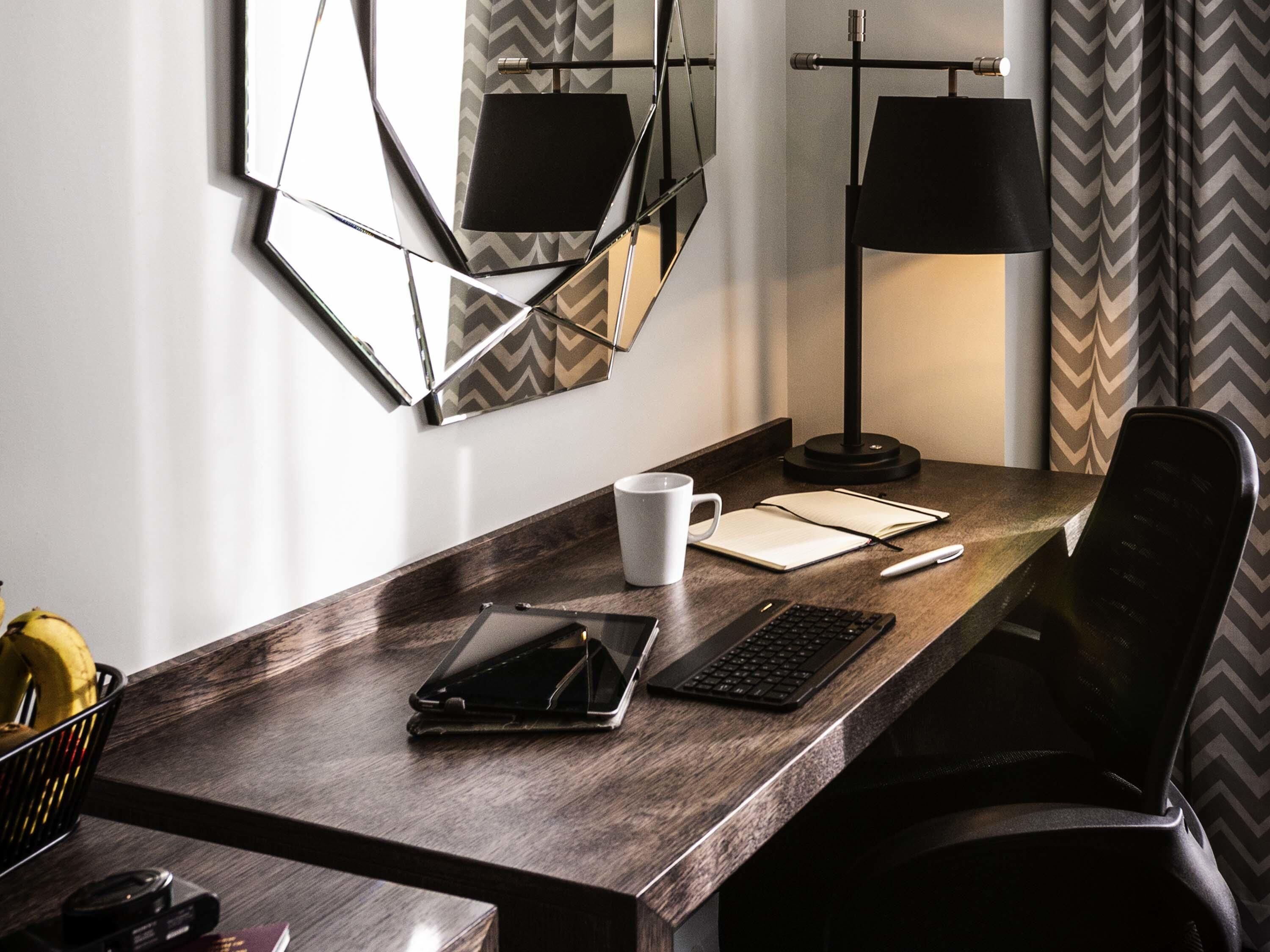 A hotel desk with a laptop, a tablet, a notebook, a pen, and a cup.
