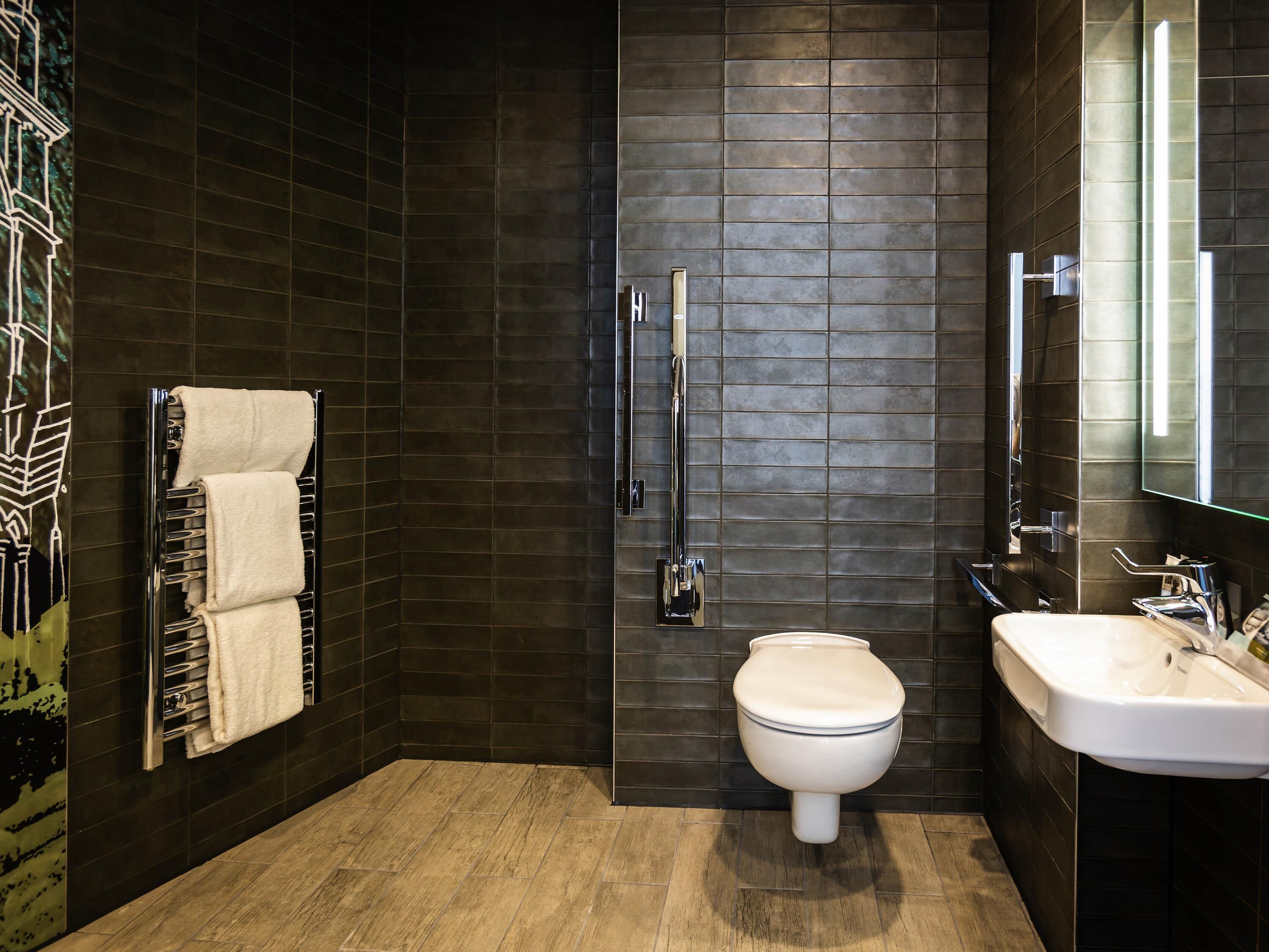 Un baño moderno con paredes de baldosas oscuras, suelo de madera, inodoro blanco, cabina de ducha de cristal y lavamanos blanco con espejo.