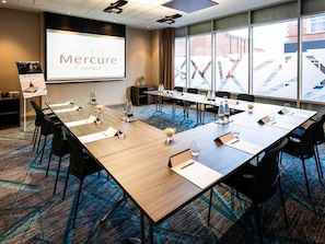 A conference room with a long table, chairs, a projector screen, and a large window with a view of buildings. - Mercure Bridgwater Hotel (Bridgwater)