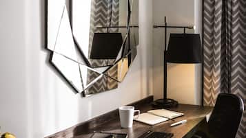 Ett modernt hotellrum med ett skrivbord i trä, en bärbar dator, en smartphone, en surfplatta, en svart stol, en spegel som hänger på väggen, en lampa och en mönstrad gardin.