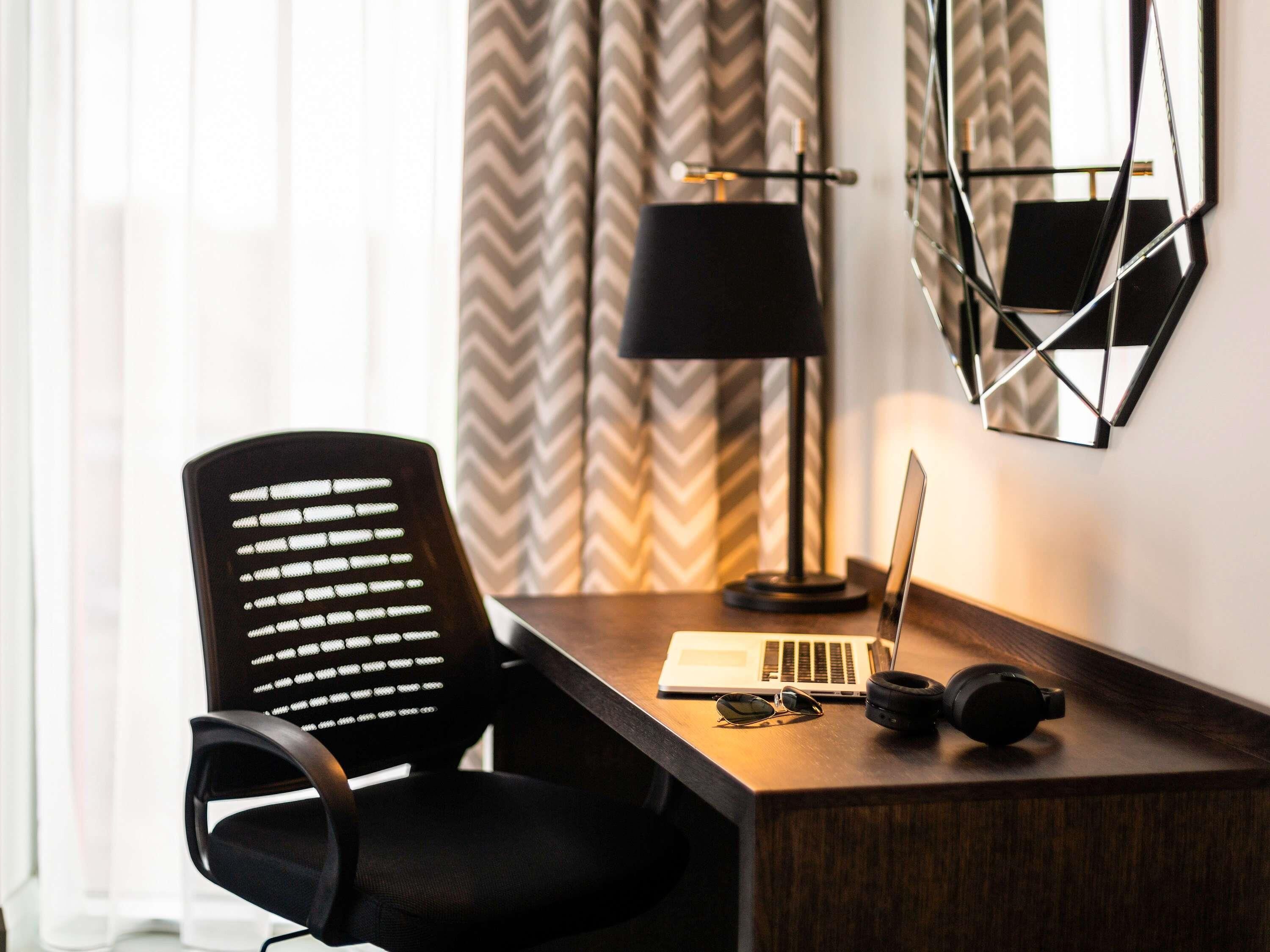A hotel room with a wooden desk, a laptop, a pair of sunglasses, and a camera on the desk.