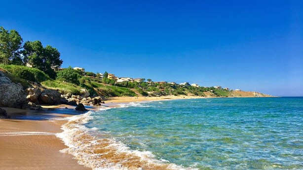 A beach with clear blue water, sandy shore, and buildings on a hillside. - Fattoria il Borghetto (Isola di Capo Rizzuto)