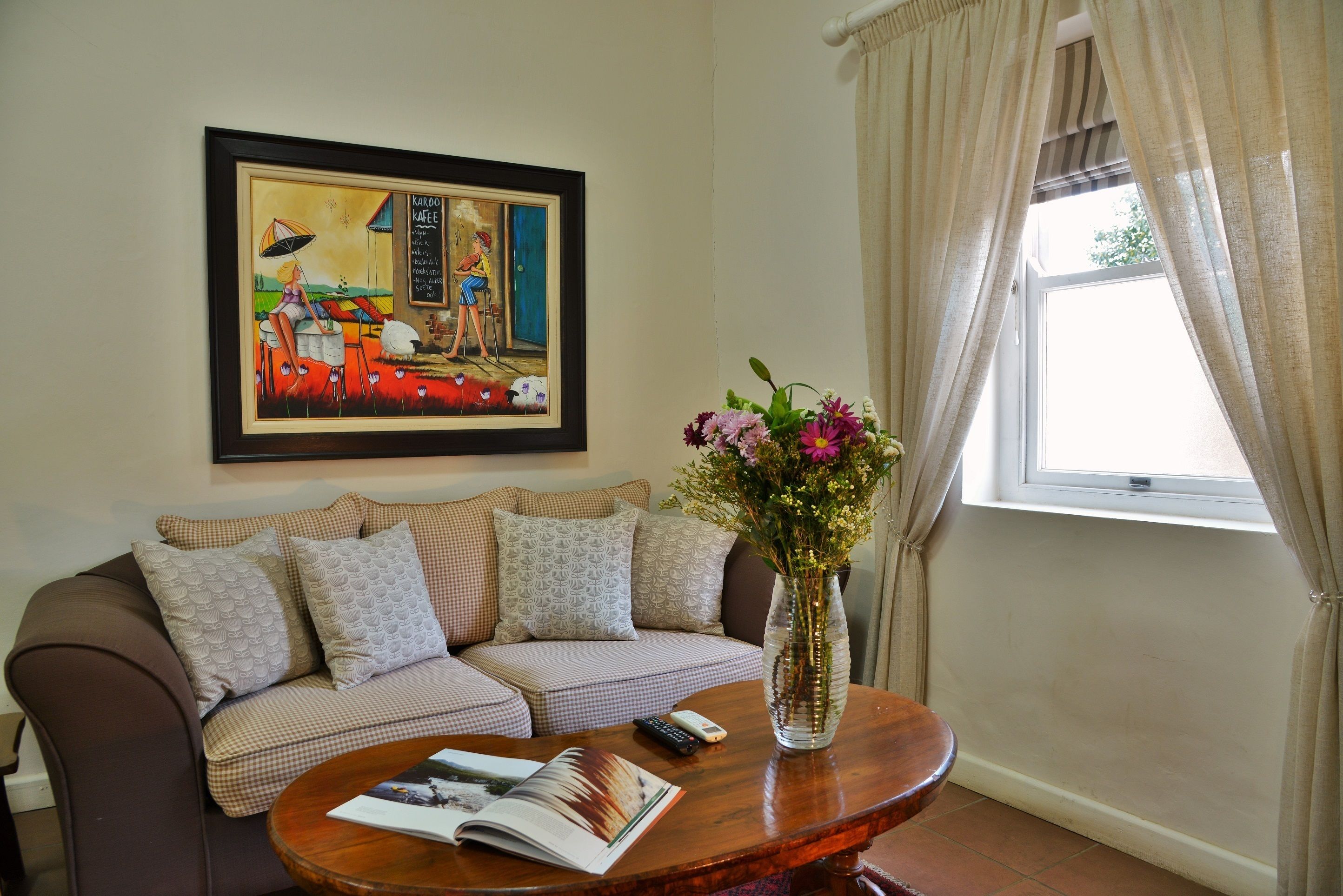 A living room with a brown sofa, a coffee table with a magazine, a framed painting, and a vase with flowers.