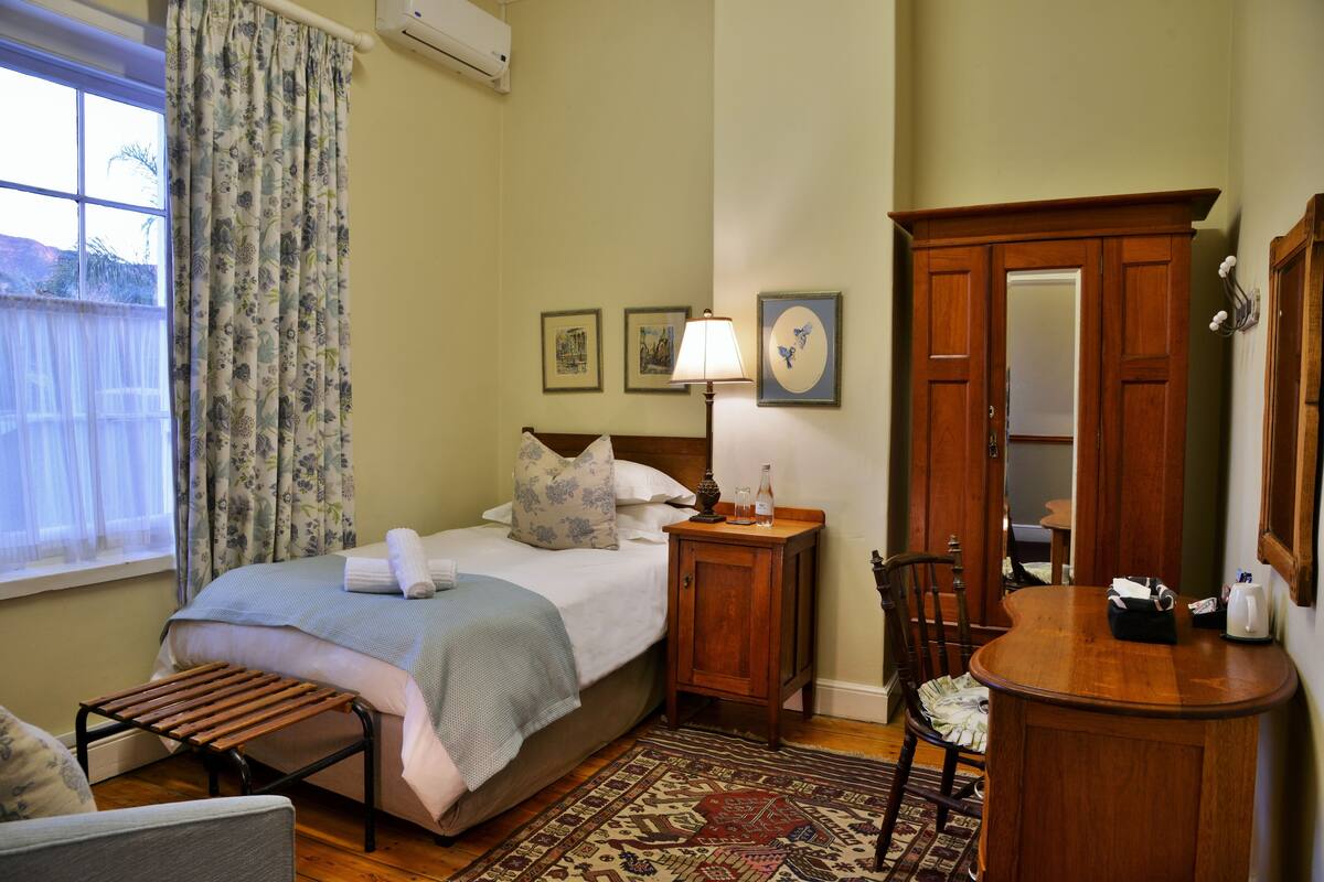 Un dormitorio con cama, un banco de madera, una silla, una mesita de noche, una lámpara, un espejo y una ventana con cortinas.