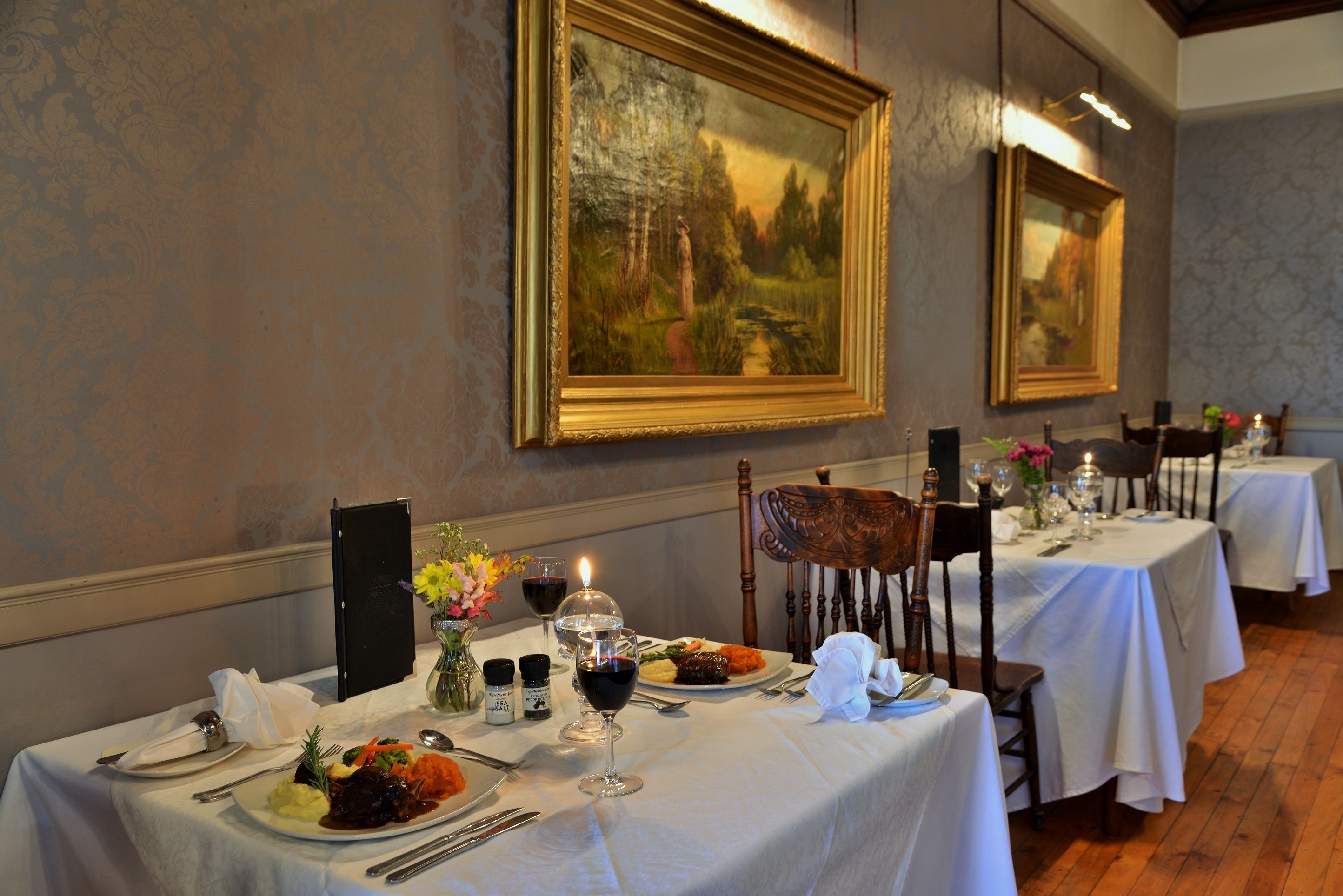A dining area with tables set for a meal, a painting on the wall, and a wooden floor.