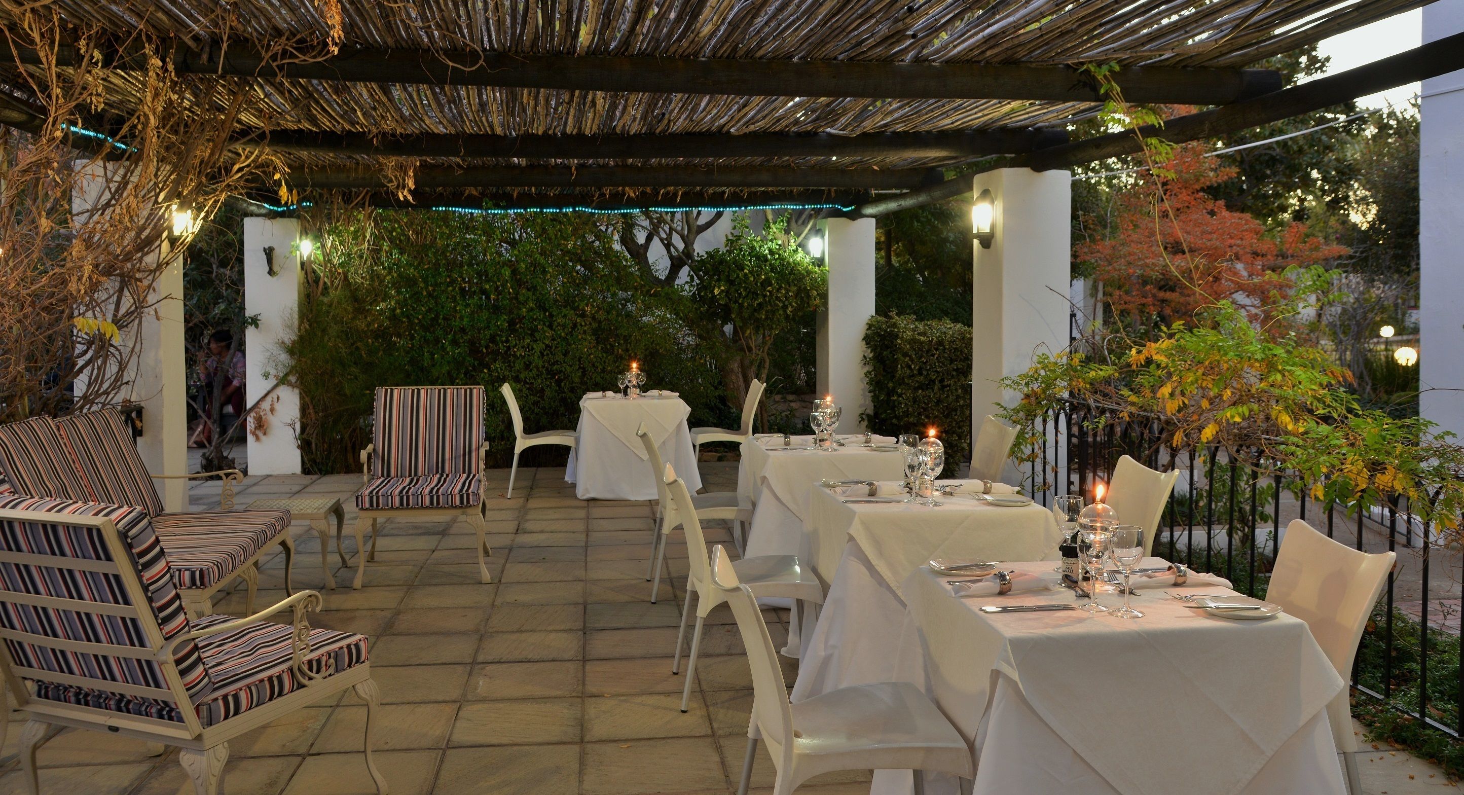 Un área de comedor al aire libre con mesas y sillas blancas, luces colgantes y una pérgola en lo alto.