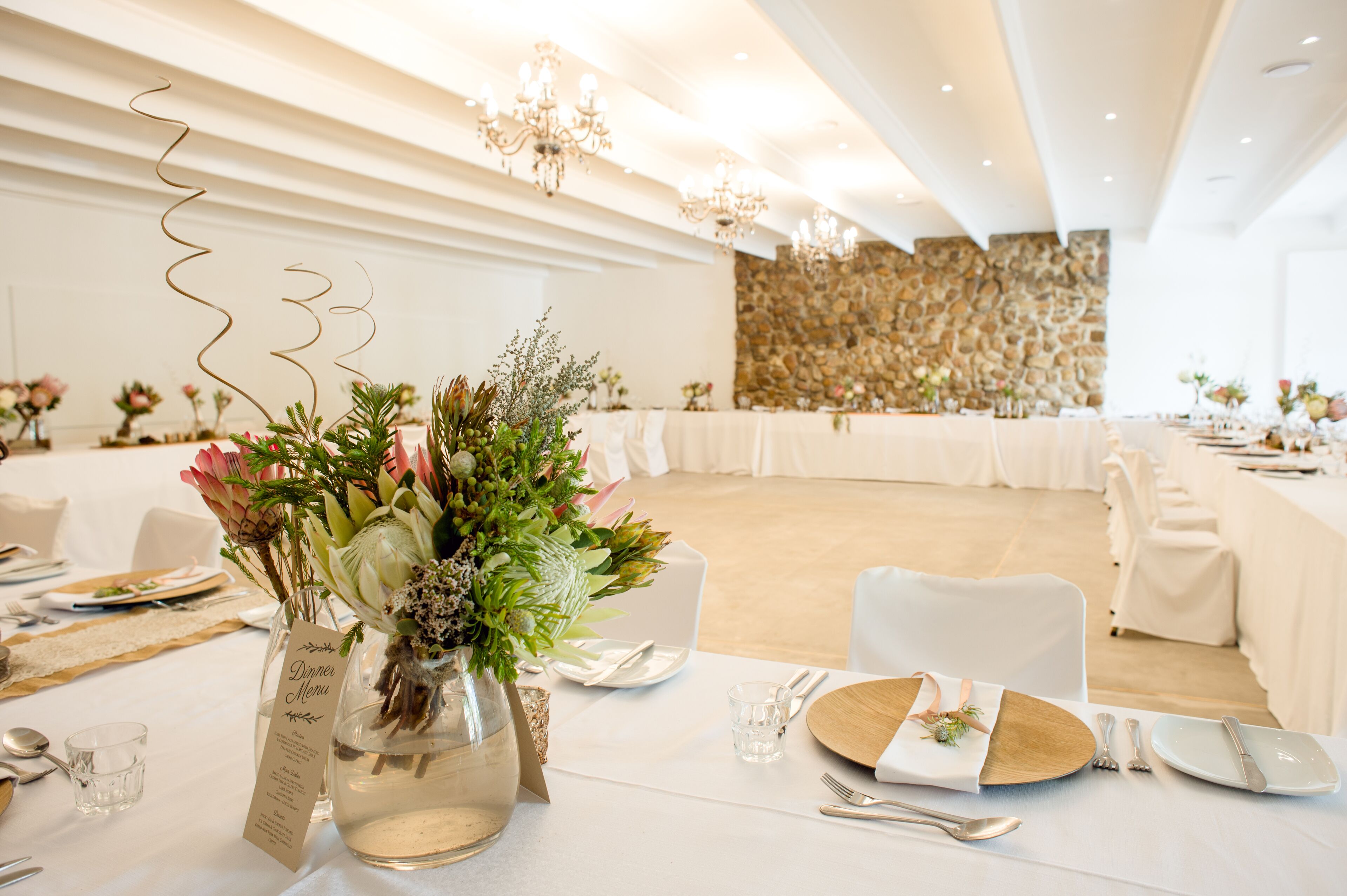 Ein gut beleuchteter Festsaal mit festlich gedeckten Tischen, weißen Tischdecken, eleganten floralen Mittelstücken und einer Wand aus Stein.