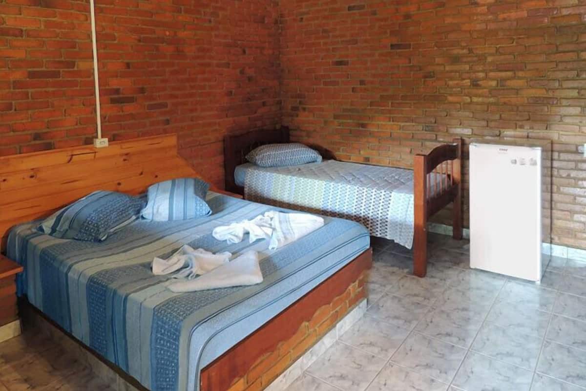 A room with a brick wall, a wooden bed, a small table, and a white refrigerator.