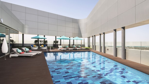 Una piscina interior moderna con sillones de descanso y sombrillas, ventanas amplias y vistas al paisaje urbano.