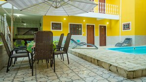 Zona junto a la piscina con sillas y una sombrilla, piscina y edificio con paredes amarillas y varias puertas.