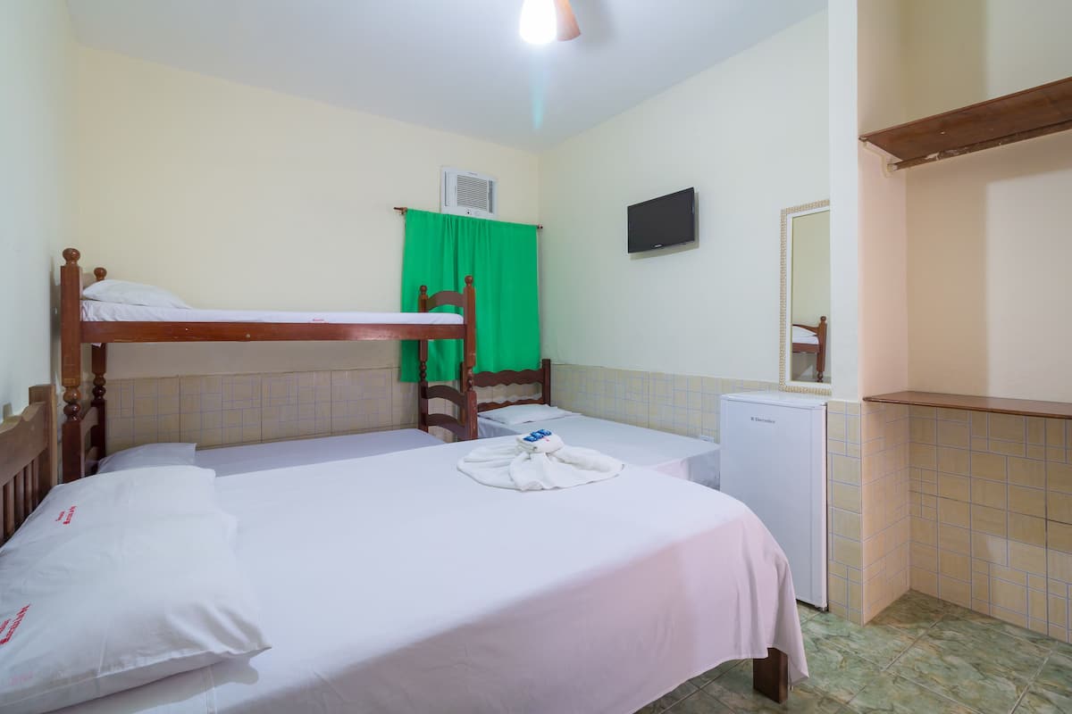 A single-bed room with a wooden headboard, a small desk with a refrigerator, a green curtain, a flat-screen TV, and a mirror on the wall.