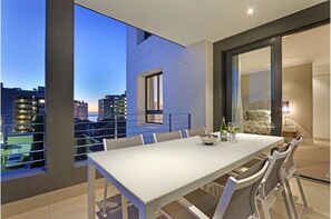Un espace de repas moderne avec une longue table et des chaises, de grandes fenêtres offrant une vue sur la ville et un balcon doté d’une balustrade.