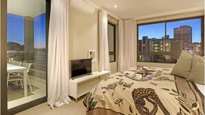 Habitación de hotel con una cama grande, televisión, balcón con mesa y sillas, y vistas a la ciudad.