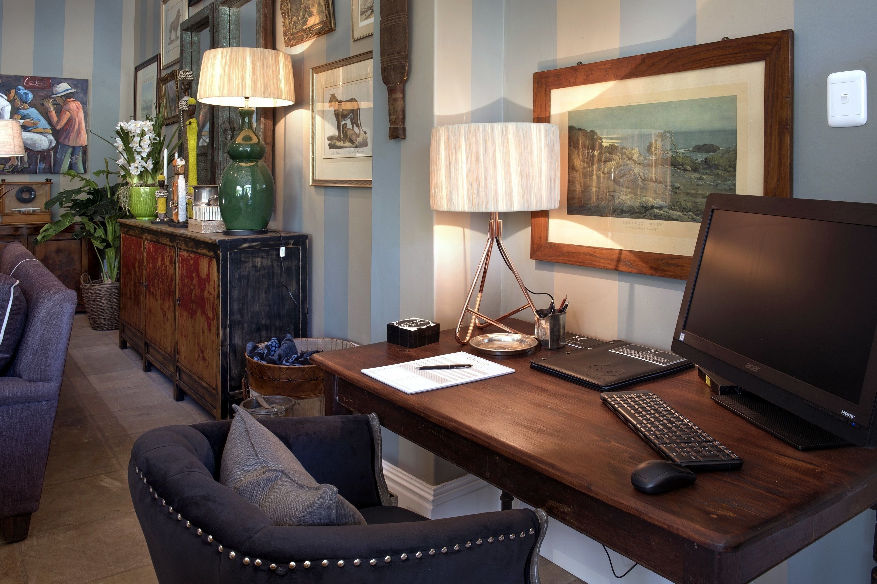 A wooden desk with a laptop, keyboard, and mouse, a lamp, framed pictures, and a couch in a room with striped walls.