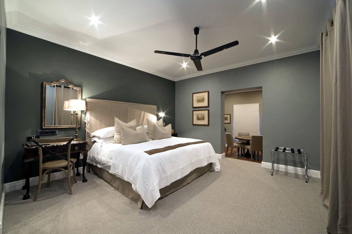 A bedroom with a large bed, a desk with a chair, a ceiling fan, and framed pictures on the wall.