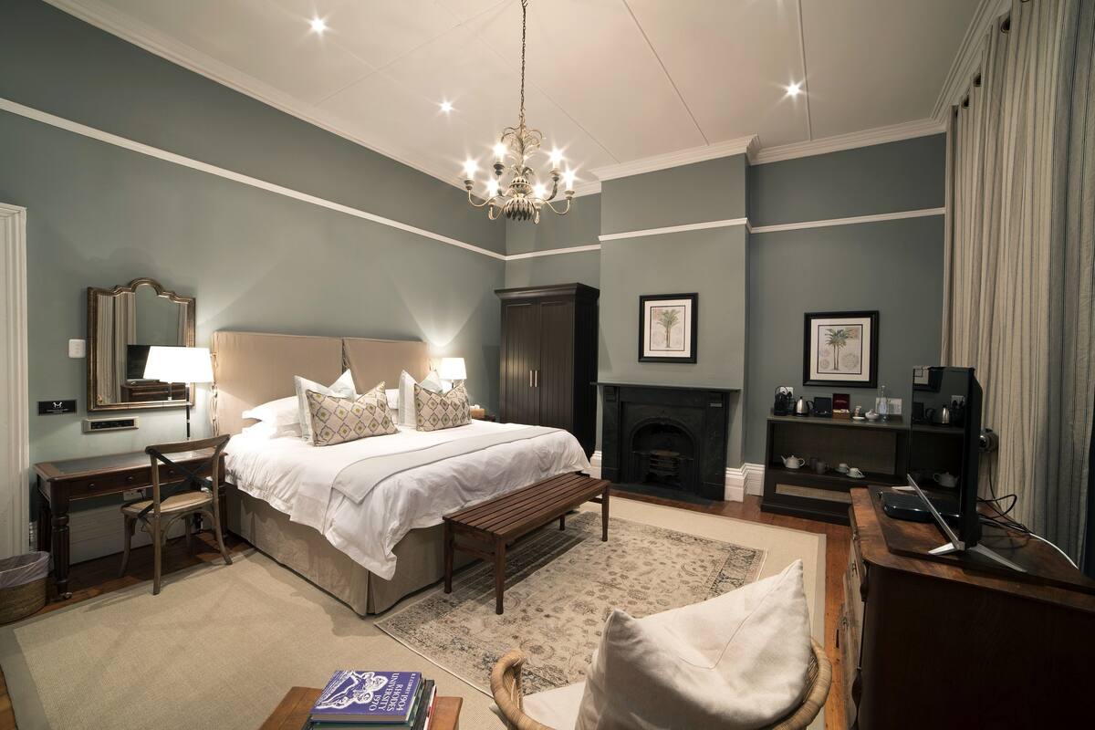 A bedroom with a large bed, a wooden bench, a desk, a chair, a fireplace, and a bookshelf.