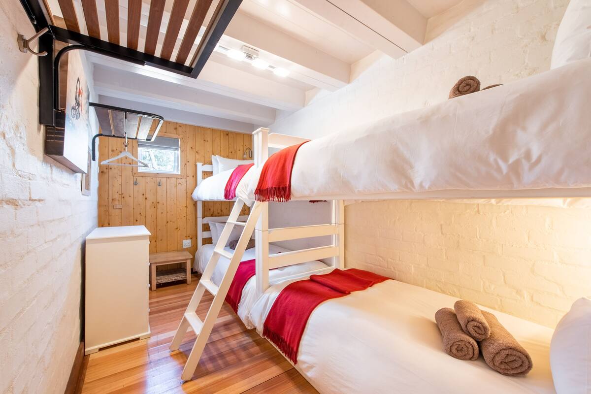 A bunk bed room with a wooden ladder, a small table, and a white wall with a window.