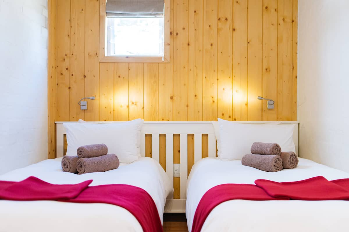 A room with two beds, wooden walls, a small window, and a red blanket on each bed.