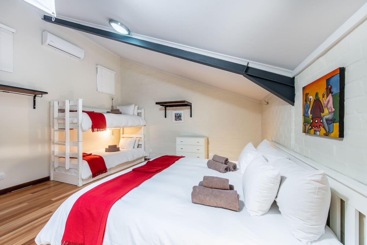A bedroom with a bunk bed, a large bed with pillows and towels, a bedside table, a nightstand, and a painting on the wall.