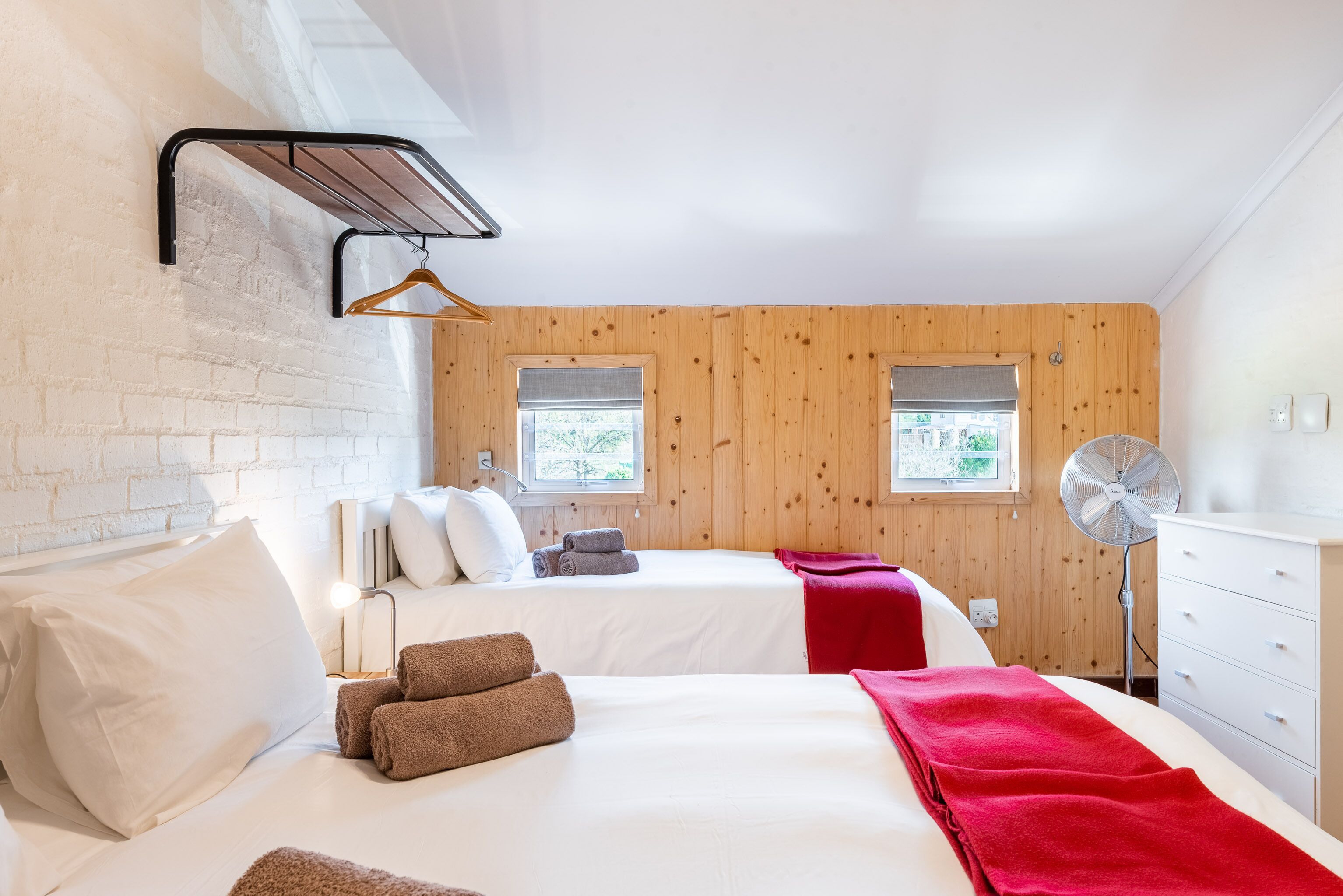 Une chambre d’hôtel avec deux lits, un mur en bois, une fenêtre, un ventilateur et une commode blanche.