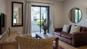 Un salon avec un canapé en cuir marron, un fauteuil en osier, une table en bois, un vase avec des plantes et une grande fenêtre avec des rideaux.