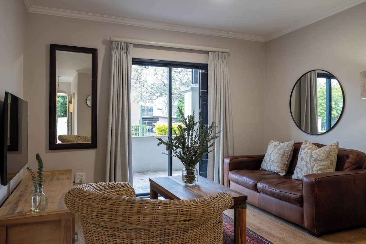 A living room with a brown leather sofa, a wicker chair, a wooden table, a vase with plants, and a large window with curtains.