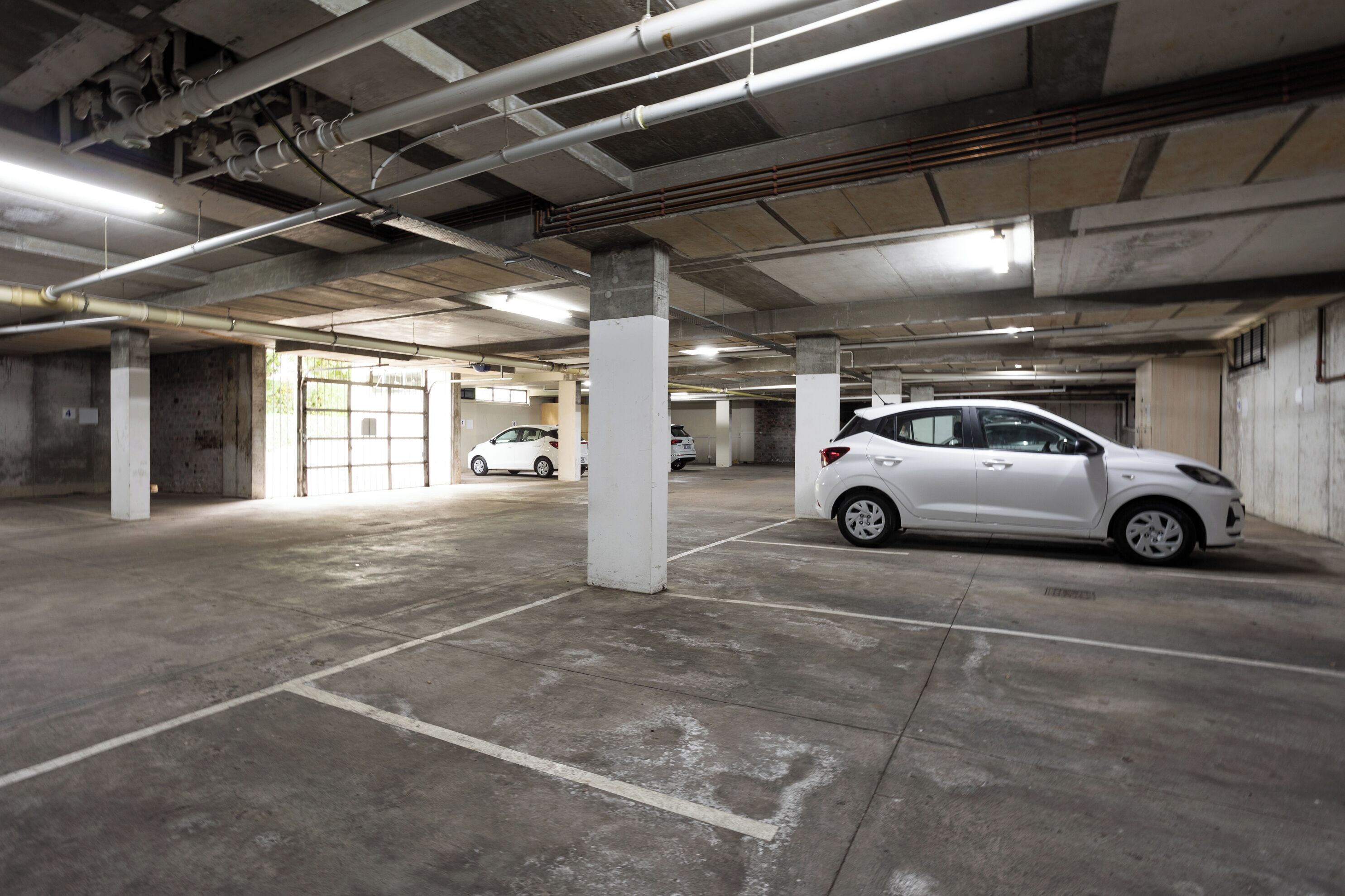 An underground parking garage with concrete pillars, fluorescent lighting, and several parked cars.