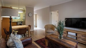 Un salon avec un canapé, une table basse et un téléviseur. On y trouve également un coin repas avec des chaises et une table. Une cuisine est visible au fond.