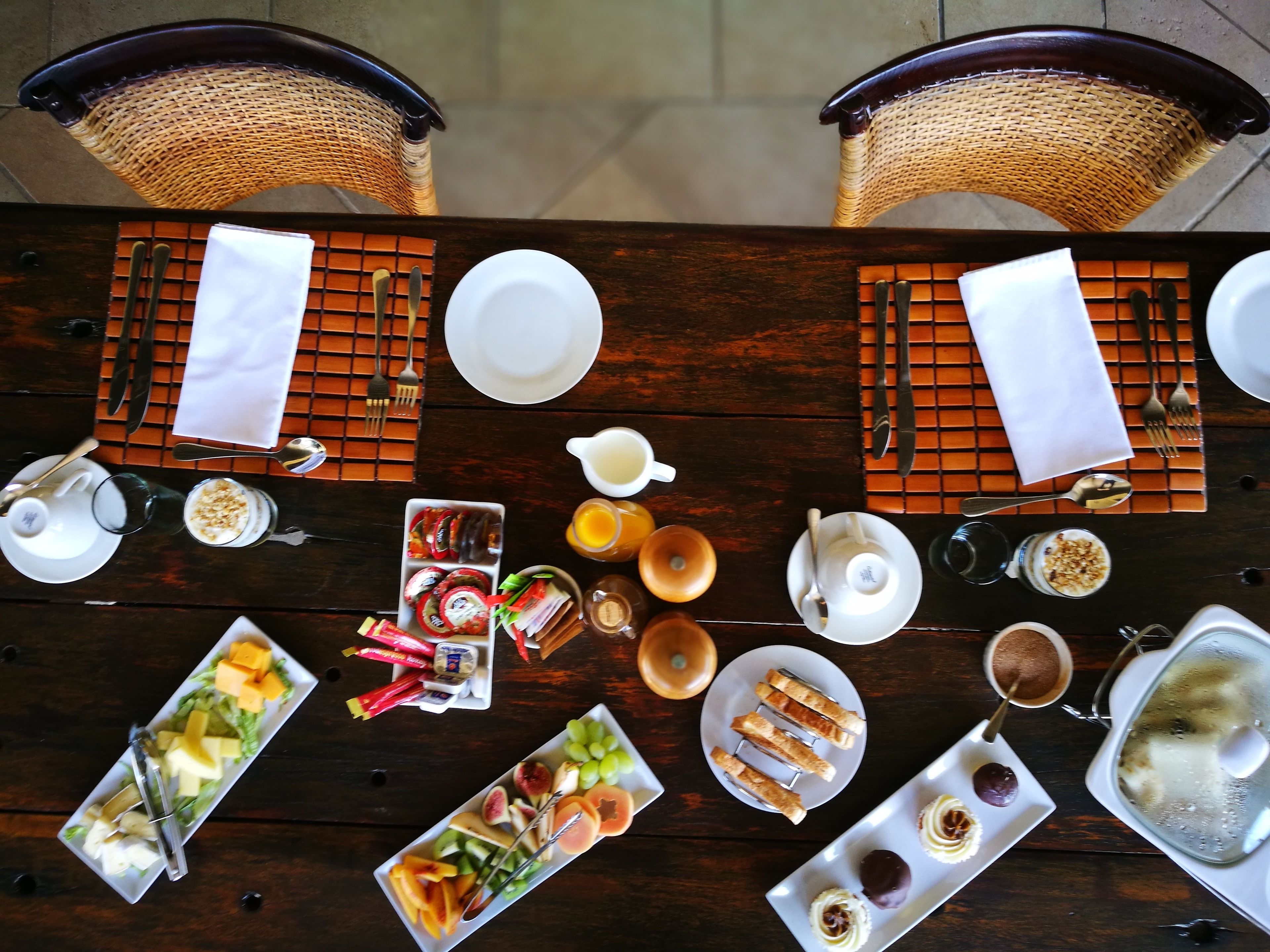 A dining table set with various dishes, including fruits, pastries, and beverages, surrounded by wicker chairs.