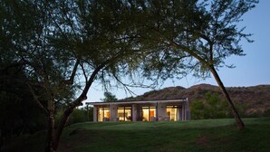 A modern house with large glass windows, surrounded by trees and a grassy hill in the background. - Wildehondekloof (Oudtshoorn)