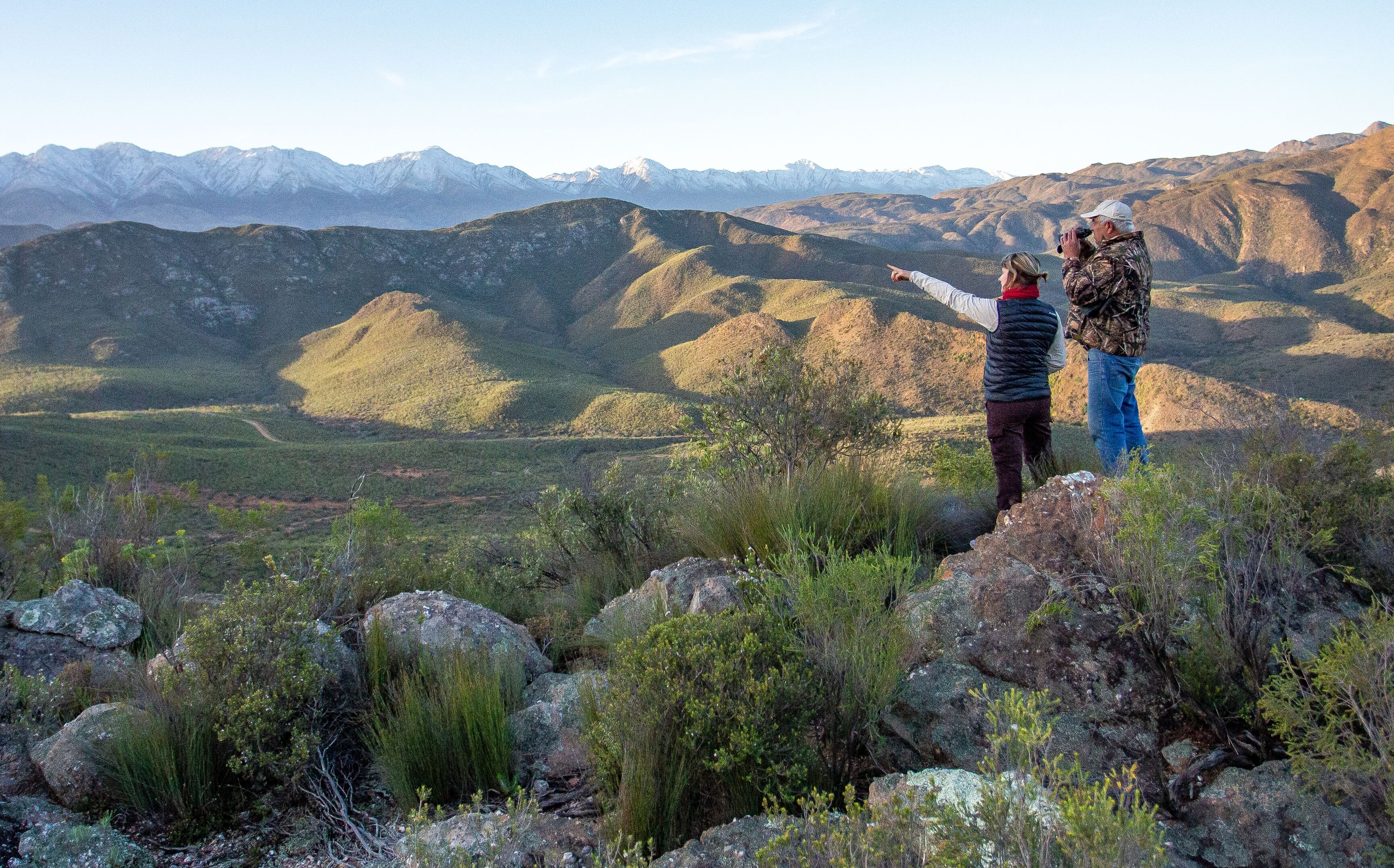 Two people standing on a rocky outcrop overlooking a mountainous landscape.