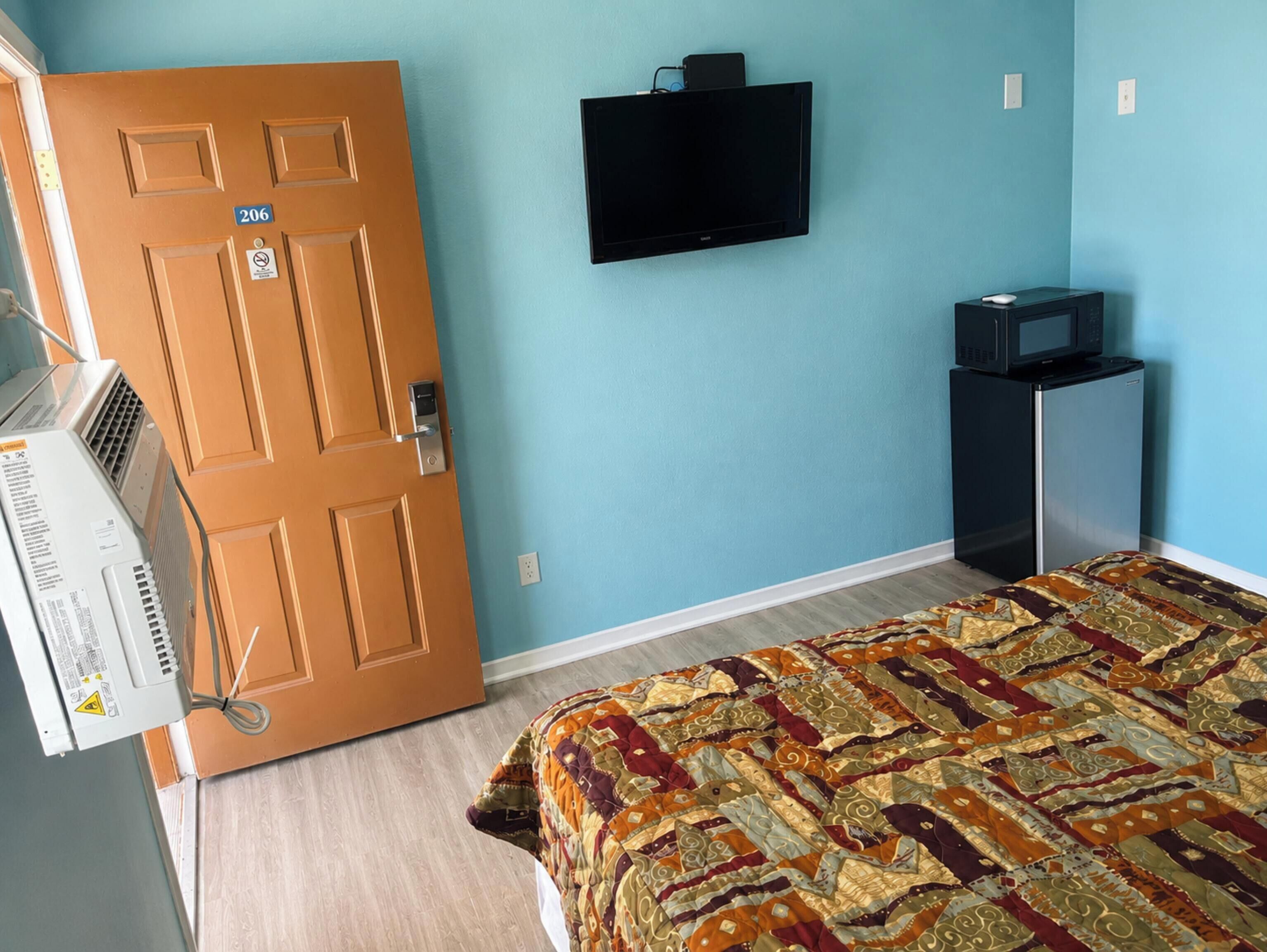 A room with a door marked 206, a flat-screen TV mounted on the wall, a microwave on a small table, and a patterned bedspread.