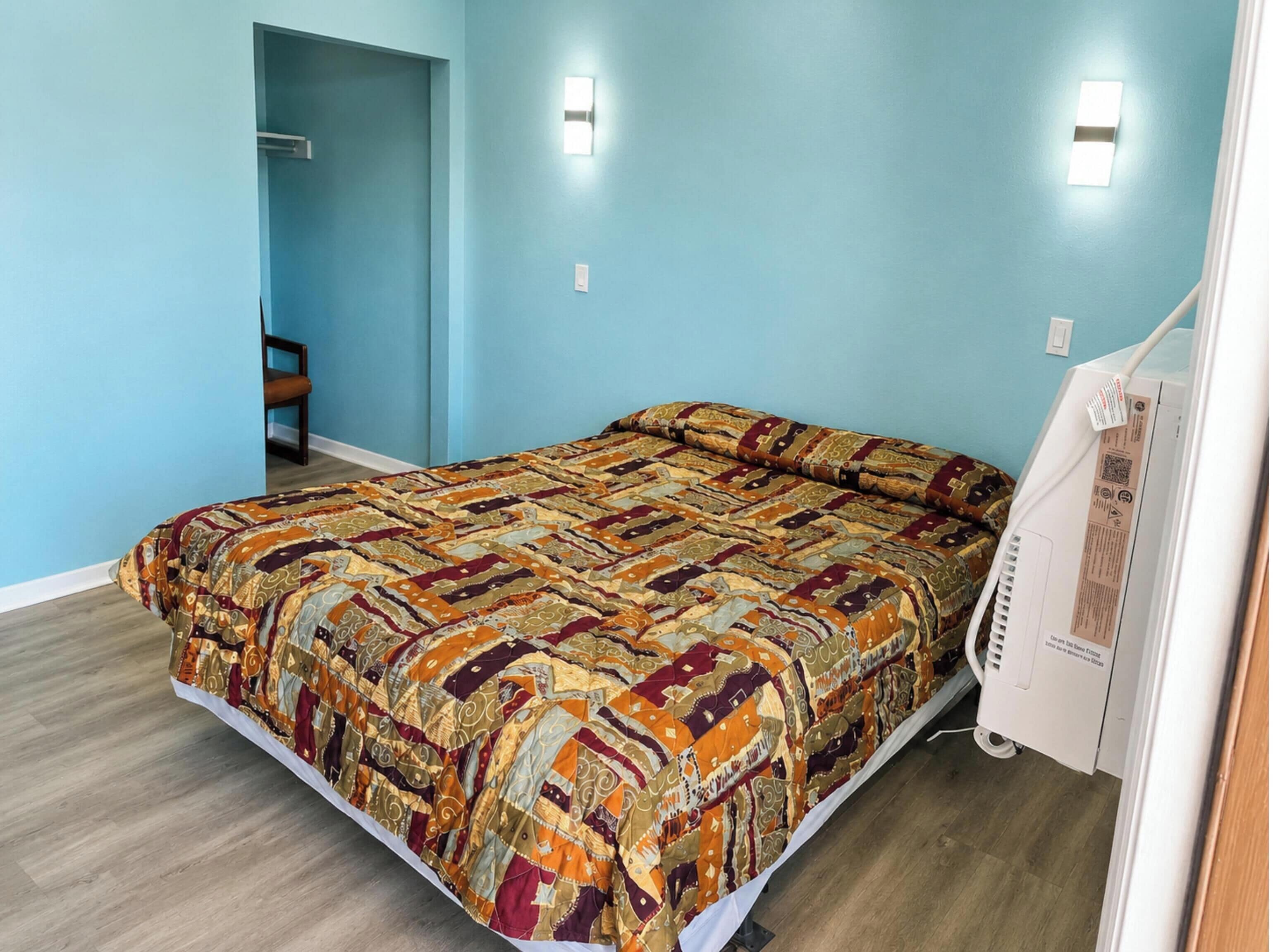 A bedroom with a patterned bedspread, a chair, a bedside table, and a wall-mounted light fixture.