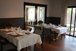 Un comedor con mesas dispuestas para cuatro comensales, cada una con mantel blanco, servilletas y cubiertos. Hay sillas alrededor de las mesas y el suelo es de madera.