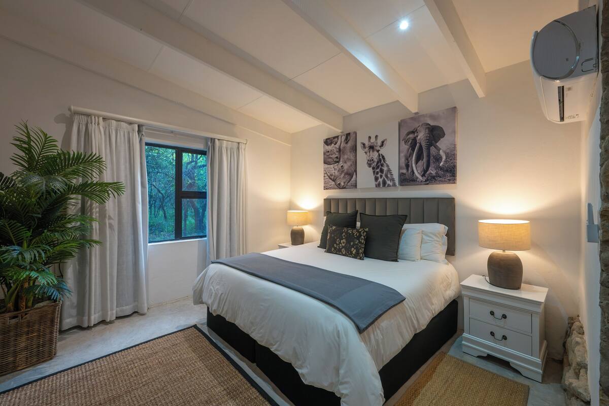 A bedroom with a large bed, two bedside tables with lamps, a window with curtains, a potted plant, and wall art.
