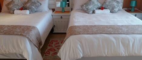 A hotel room with two beds, each with white linens and patterned pillows. There are bedside tables with lamps and a mirror above the beds.