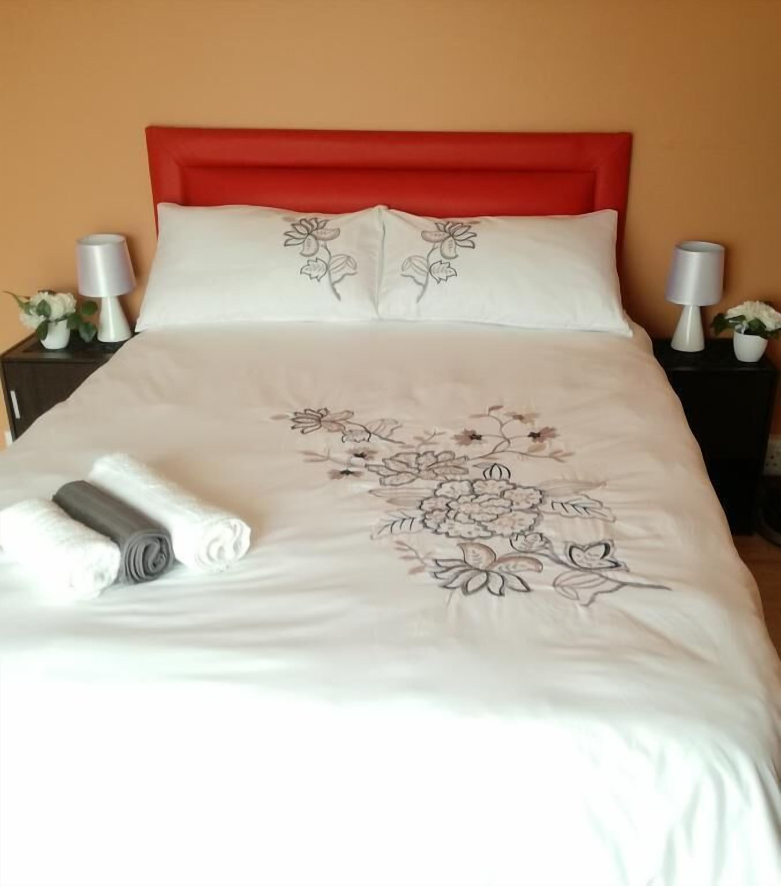 A neatly made bed with a red headboard, white pillows, and a floral bedspread. There are two bedside tables with lamps and small potted plants.