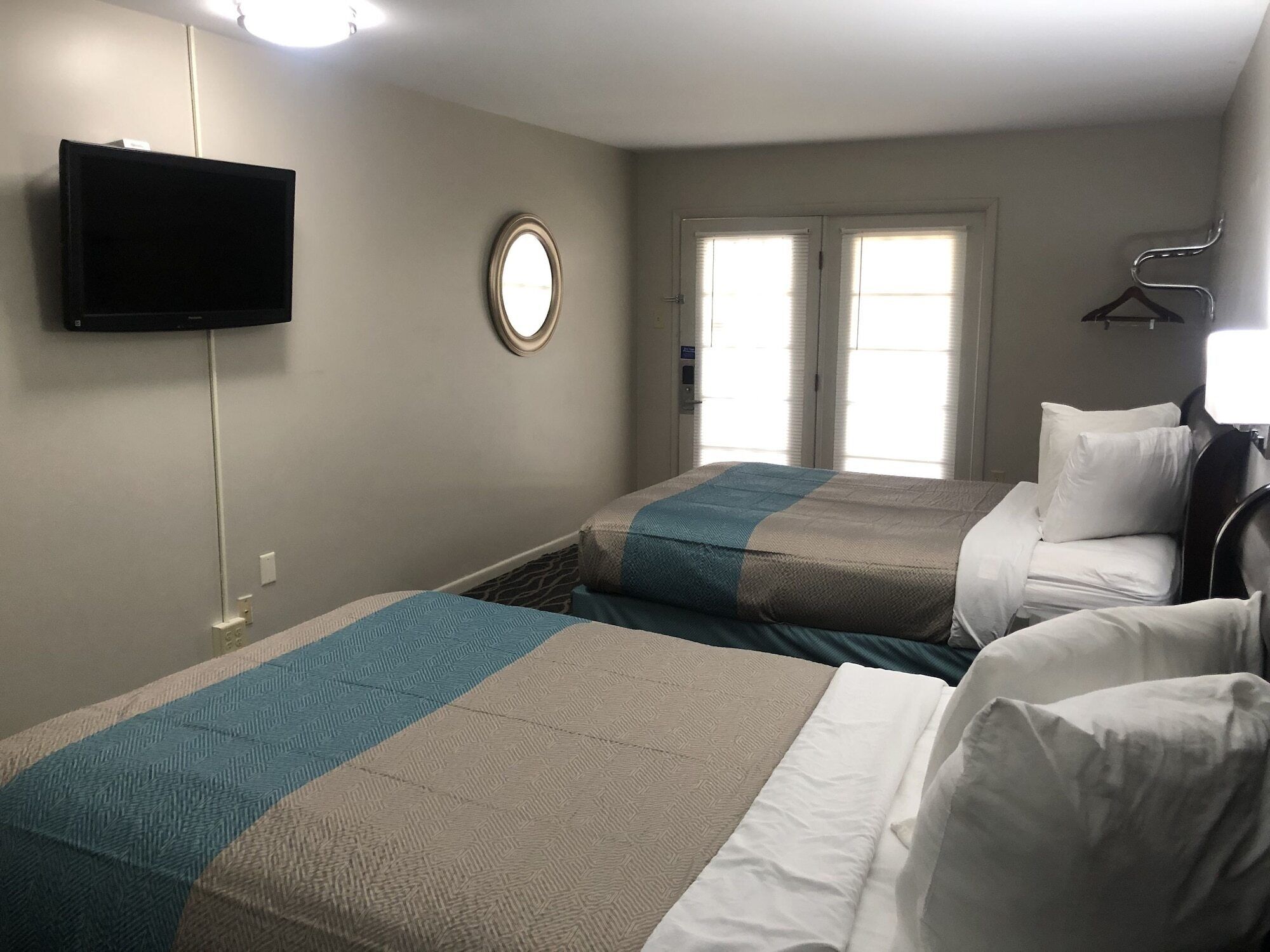 Habitación de hotel con dos camas, un televisor montado en la pared, un espejo redondo y una ventana con persianas.