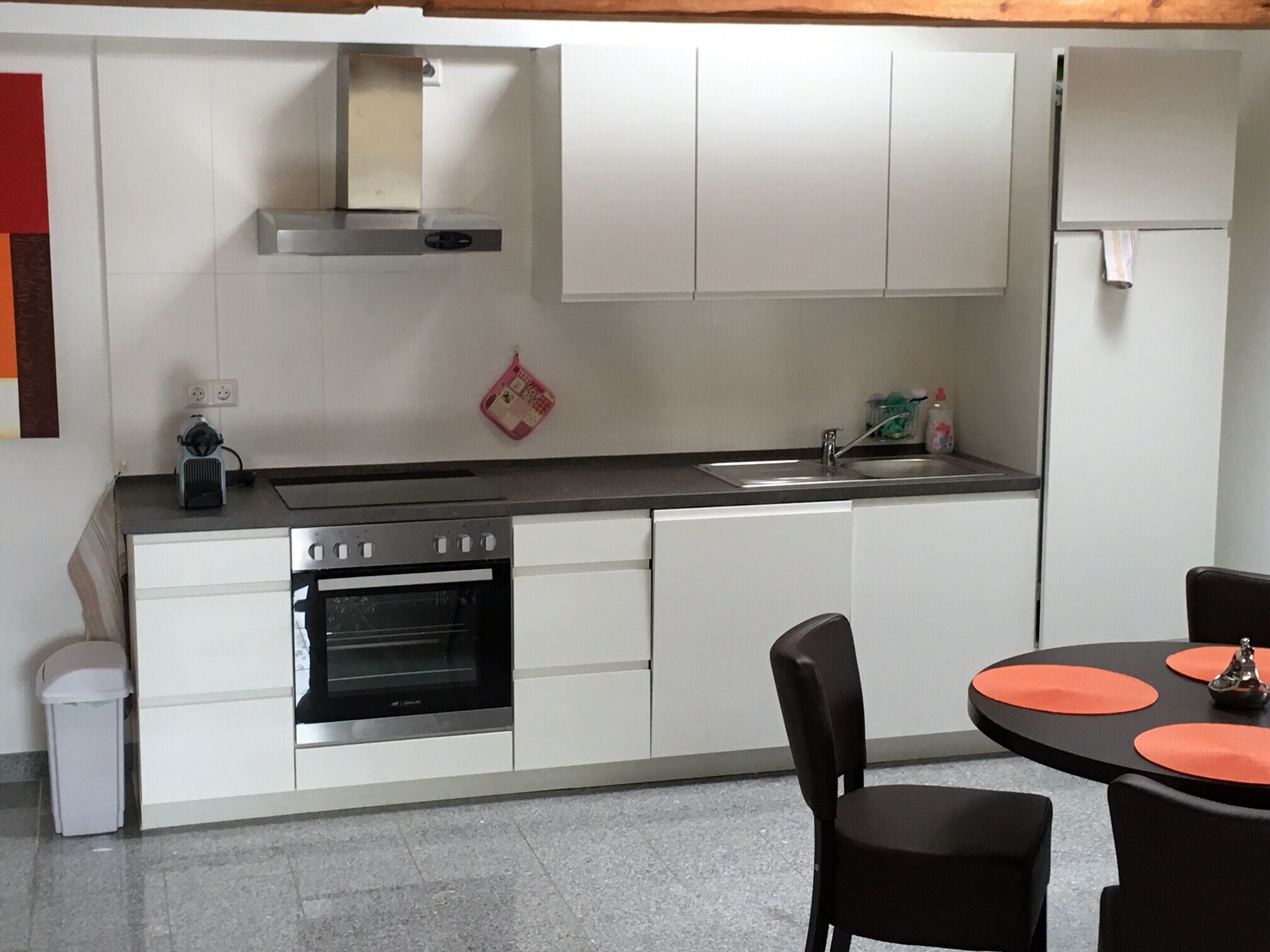 Eine moderne Küche mit weißen Schränken, einer Edelstahl-Dunstabzugshaube, einem Einbauofen, einer Spüle und einem Essbereich mit schwarzen Stühlen und orangefarbenen Platzsets.
