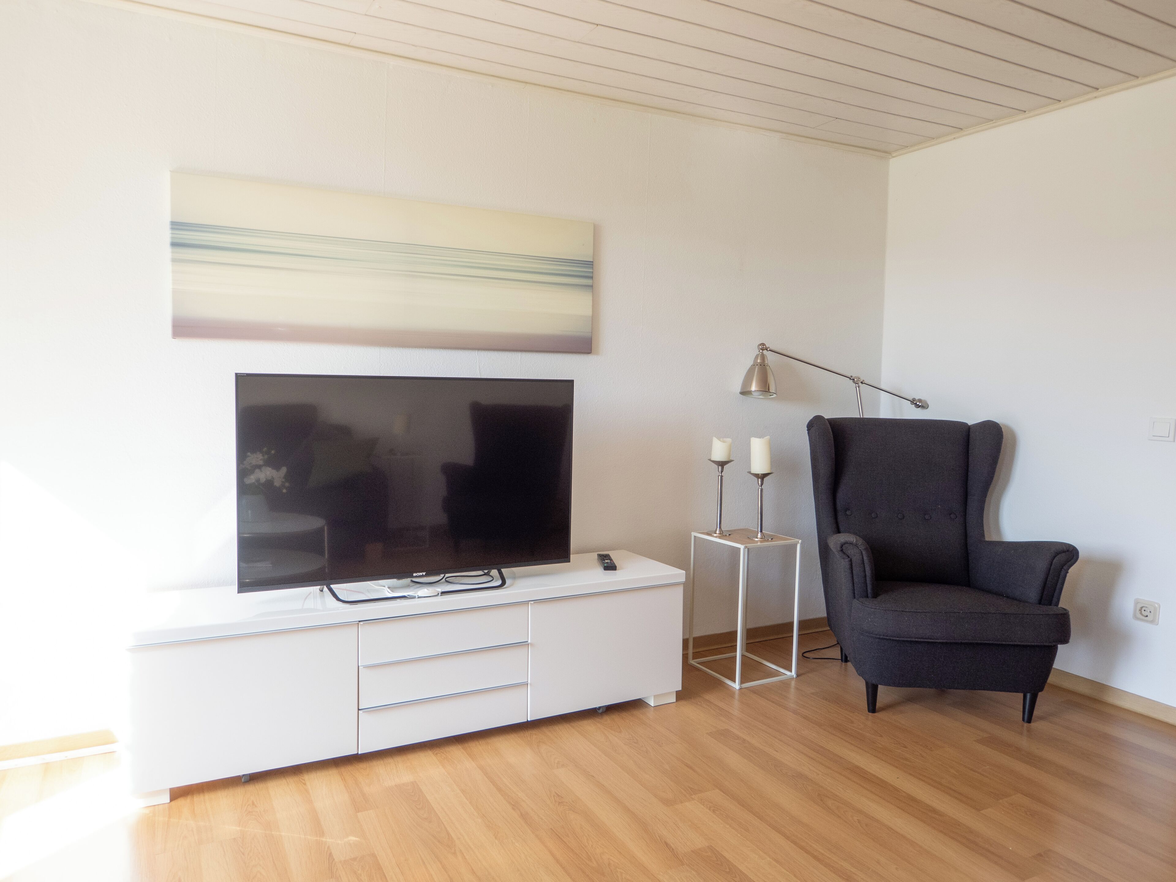Una sala de estar moderna con un televisor de pantalla plana sobre un mueble blanco, un sillón oscuro y un cuadro minimalista en la pared.